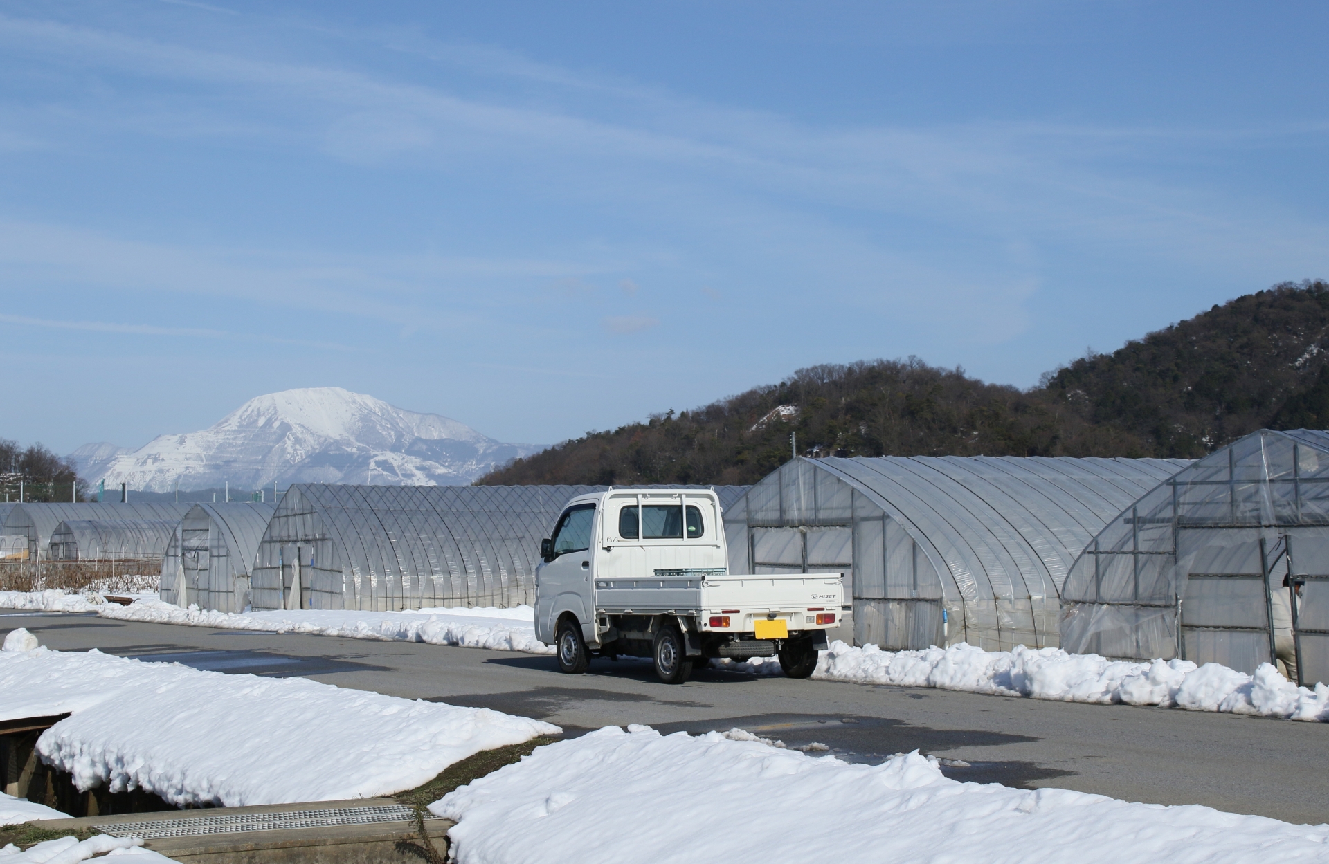 滋賀県の名峰、伊吹山が見える畑のビニールハウスの前に駐車している軽トラックと雪景色